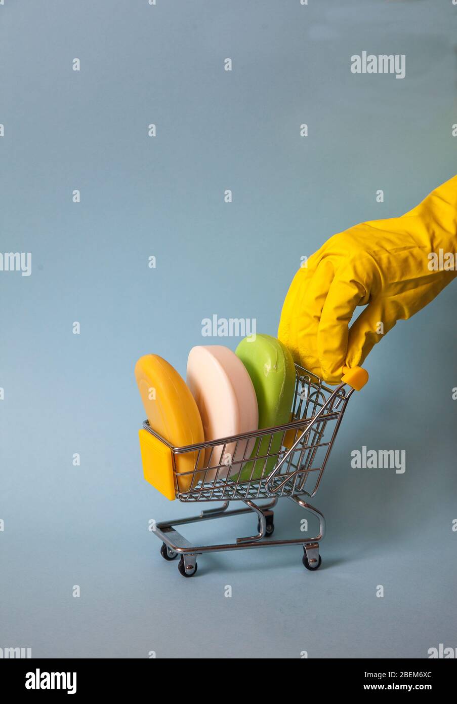 Una mano femenina en un guante amarillo enrolla un carro de supermercado con trozos de jabón sobre un fondo azul. Higiene. Desinfección. Covid 19. Espas de la copia. Foto de stock