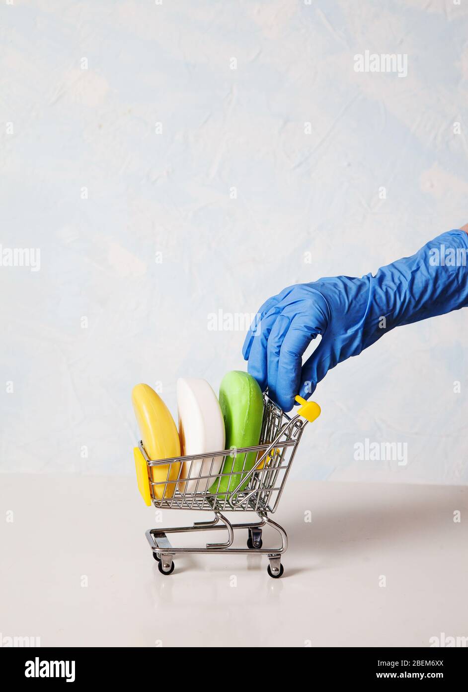 Una mano femenina en un guante azul enrolla un carro de supermercado con trozos de jabón sobre un fondo blanco. Higiene. Desinfección. Covid 19. Espas de la copia. Foto de stock