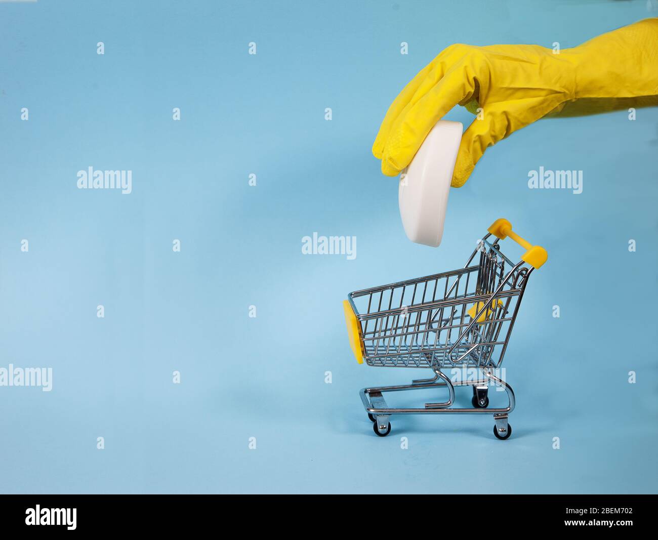 La mano femenina en un guante amarillo sumerge una barra de jabón en un carrito de supermercado sobre un fondo azul. Desinfección, Coronvirus. Covid 19. Copiar espas. Foto de stock
