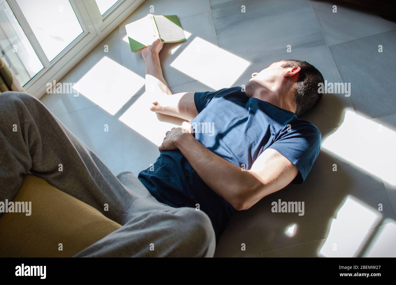 Un joven aburrido se aloja en casa. Hombre acostado en el suelo leyendo y disfrutando del sol de la ventana. Concepto de estancia en casa, aburrimiento, libertad, soledad. Foto de stock