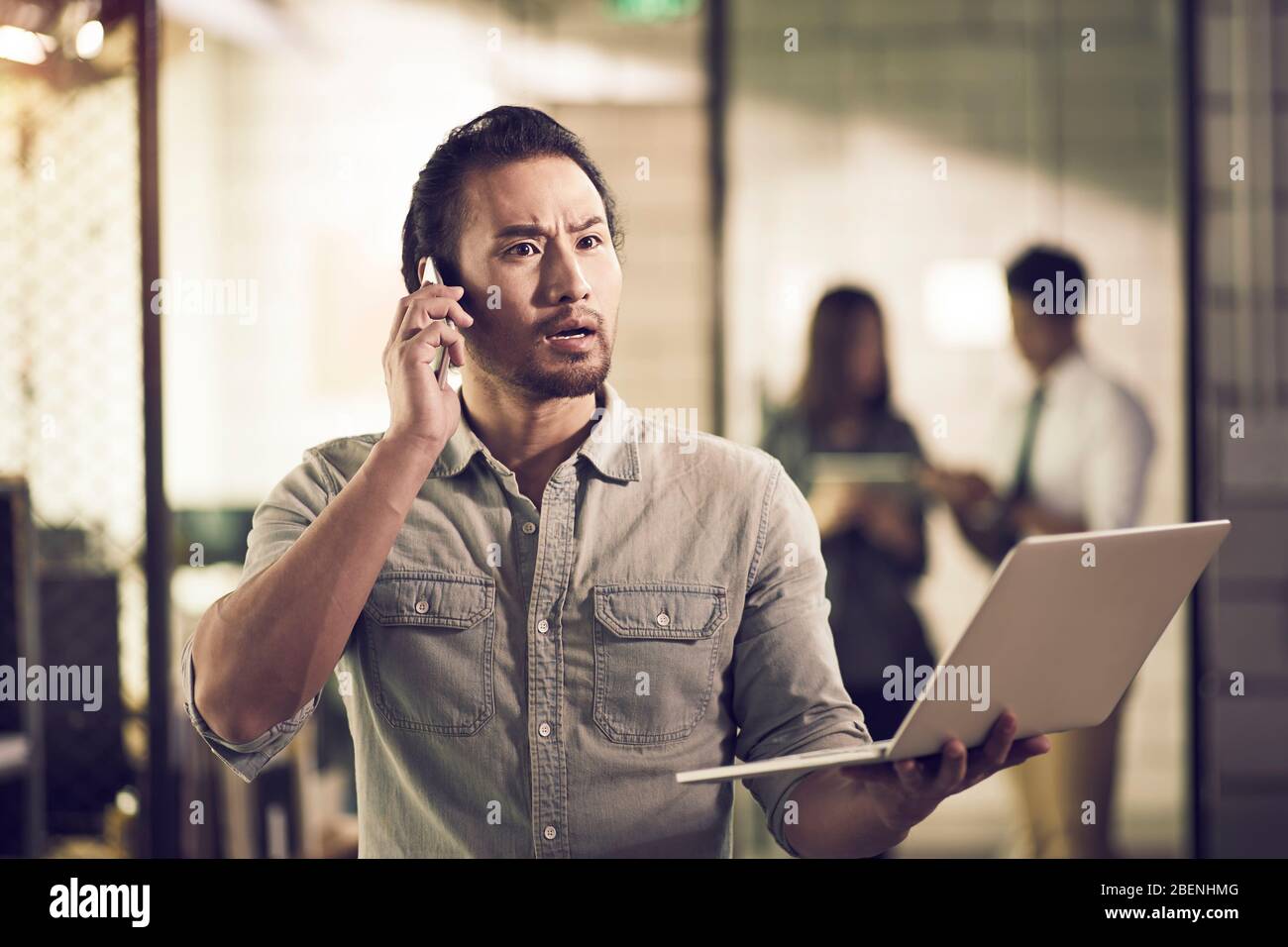 empresario asiático sosteniendo un ordenador portátil haciendo una llamada usando el teléfono celular y conmocionado por malas noticias Foto de stock