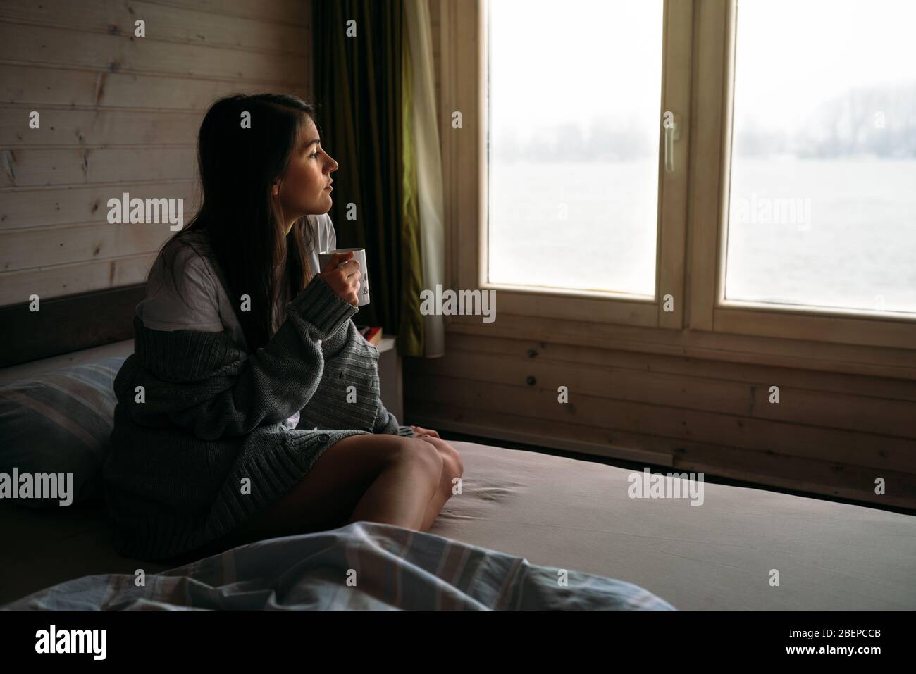 Mujer joven que se aloja en casa bebiendo café/té, mirando a través de la ventana.comenzando el día, ritual de la mañana.Quaratine aislamiento de uno mismo Concept.Proven wom Foto de stock