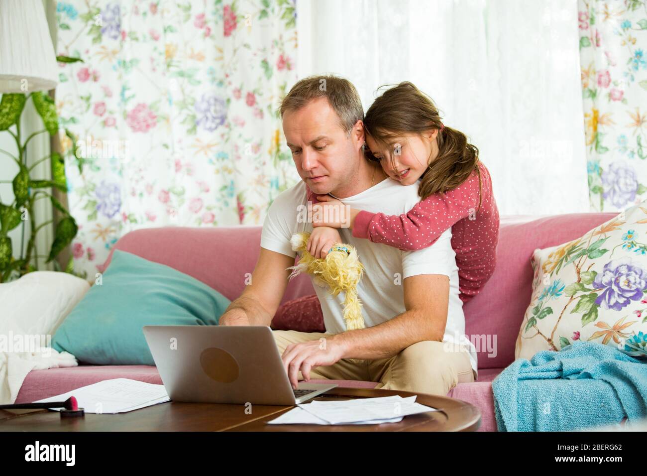 Niño jugando y perturbando a padre trabajando a distancia de casa. Hombre sentado en el sofá con ordenador portátil. Familia pasando tiempo juntos en el interior. Foto de stock