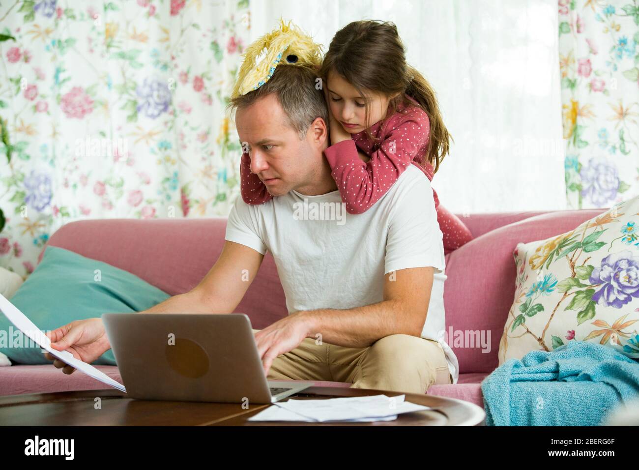 Niño jugando y perturbando a padre trabajando a distancia de casa. Hombre sentado en el sofá con ordenador portátil. Familia pasando tiempo juntos en el interior. Foto de stock