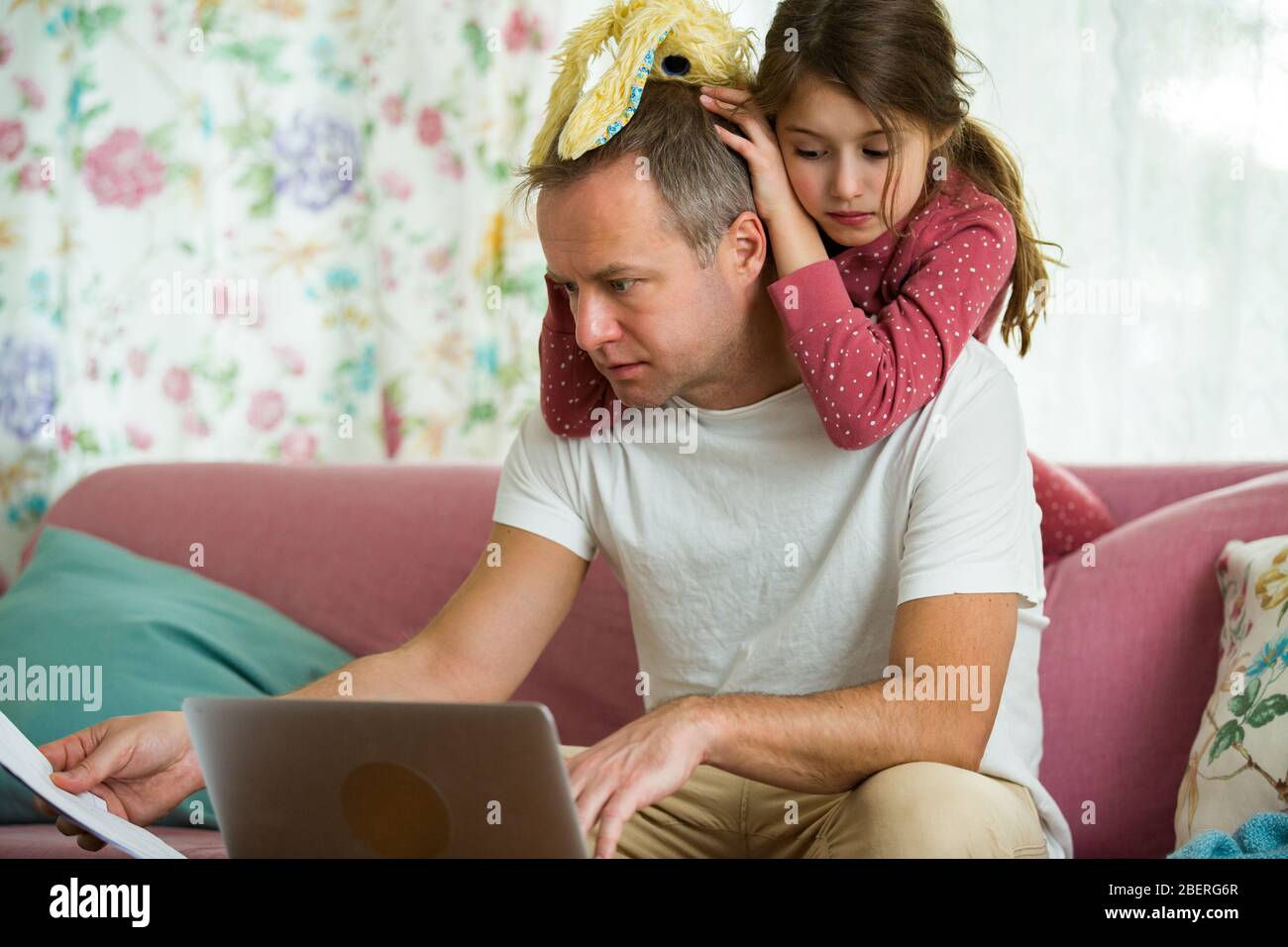 Niño jugando y perturbando a padre trabajando a distancia de casa. Hombre sentado en el sofá con ordenador portátil. Familia pasando tiempo juntos en el interior. Foto de stock