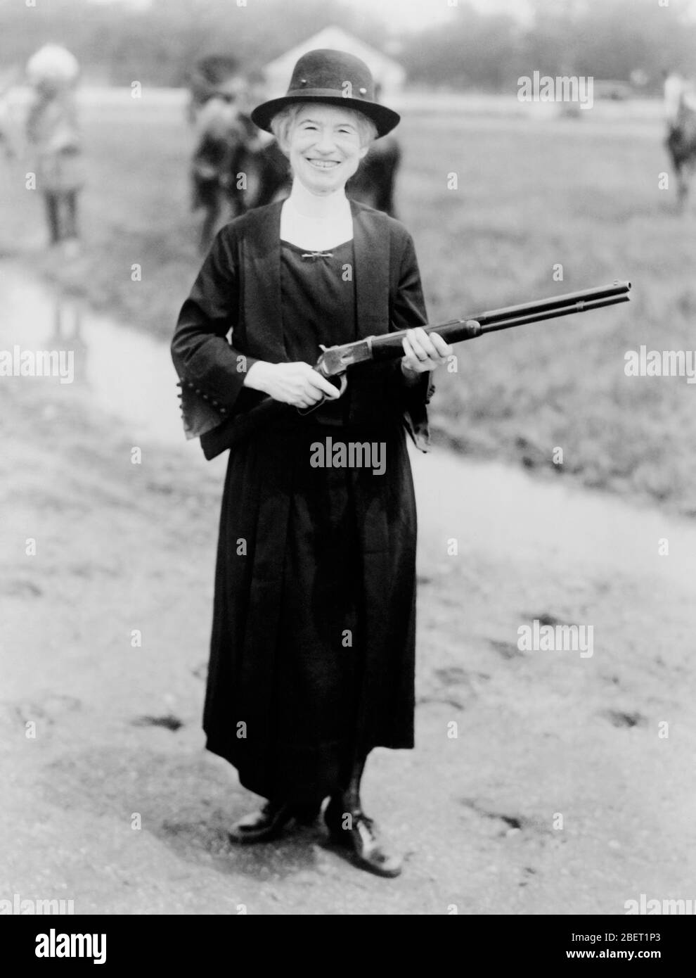 Annie Oakley de pie en un sombrero negro y vestido mientras sostiene un rifle, 1922. Foto de stock