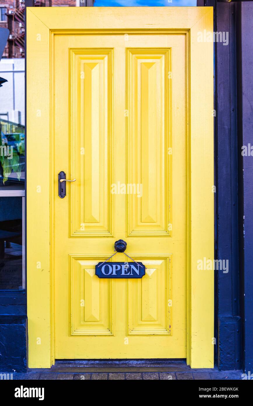 Puerta de madera con paneles de color amarillo brillante y "ABIERTA" colgada de un pomo de puerta de latón que crea un llamativo contraste en una calle Launceston de Tasmania. Foto de stock