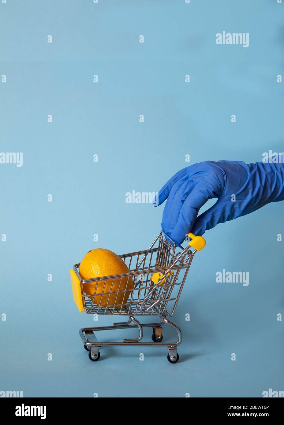 Mano femenina en un guante de goma azul enrolla un carrito de supermercado con limón sobre un fondo azul. Protección. Higiene. Covid 19 Foto de stock