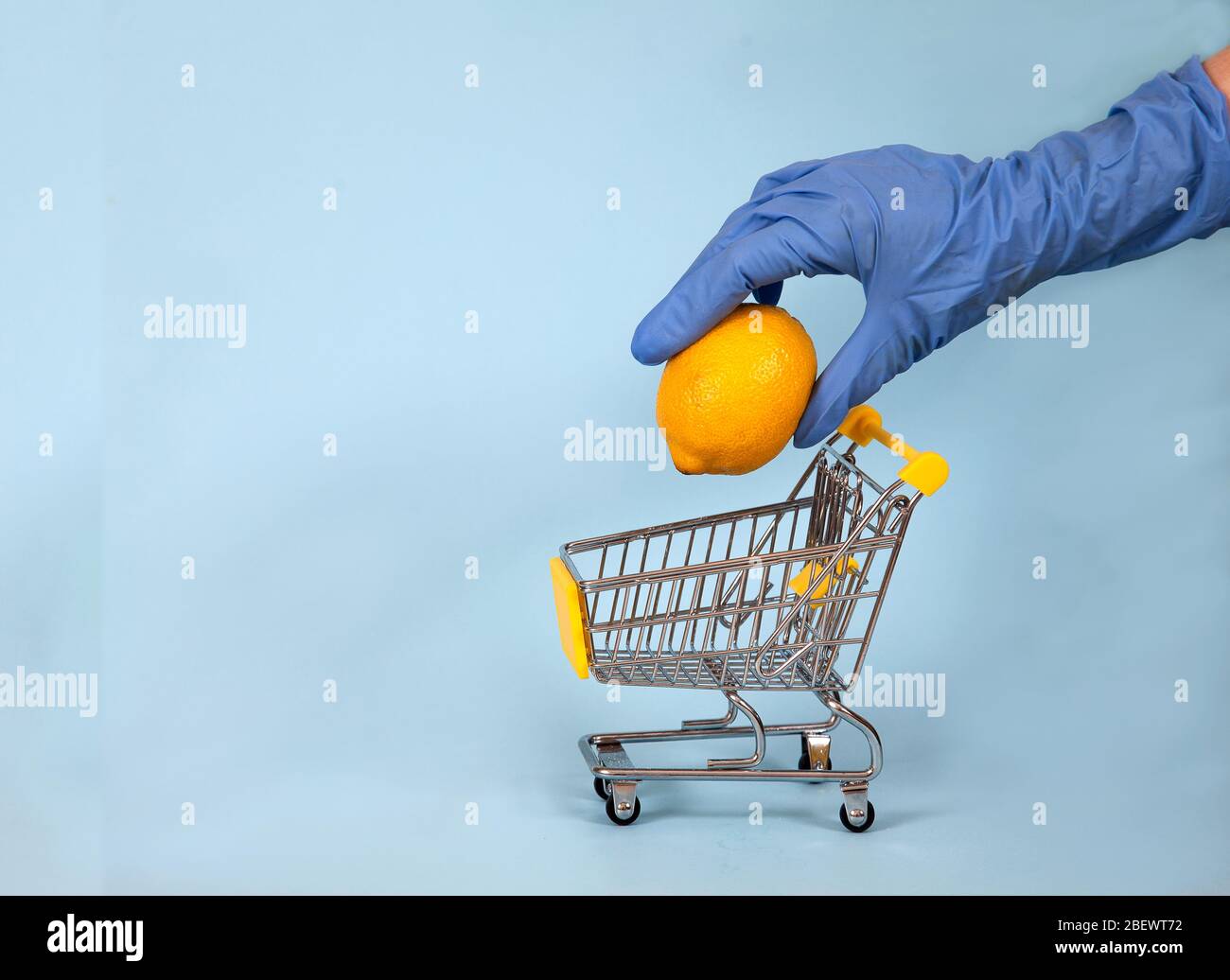 Mano femenina en un guante de goma azul baja un limón en un carrito de supermercado sobre un fondo azul. Protección. Higiene. Salud. Copiar spaes. Covid 19. Foto de stock