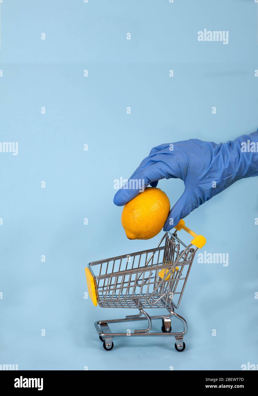 Mano femenina en un guante de goma azul baja un limón en un carrito de supermercado sobre un fondo azul. Protección. Higiene. Salud. Copiar spaes. Covid 19. Foto de stock