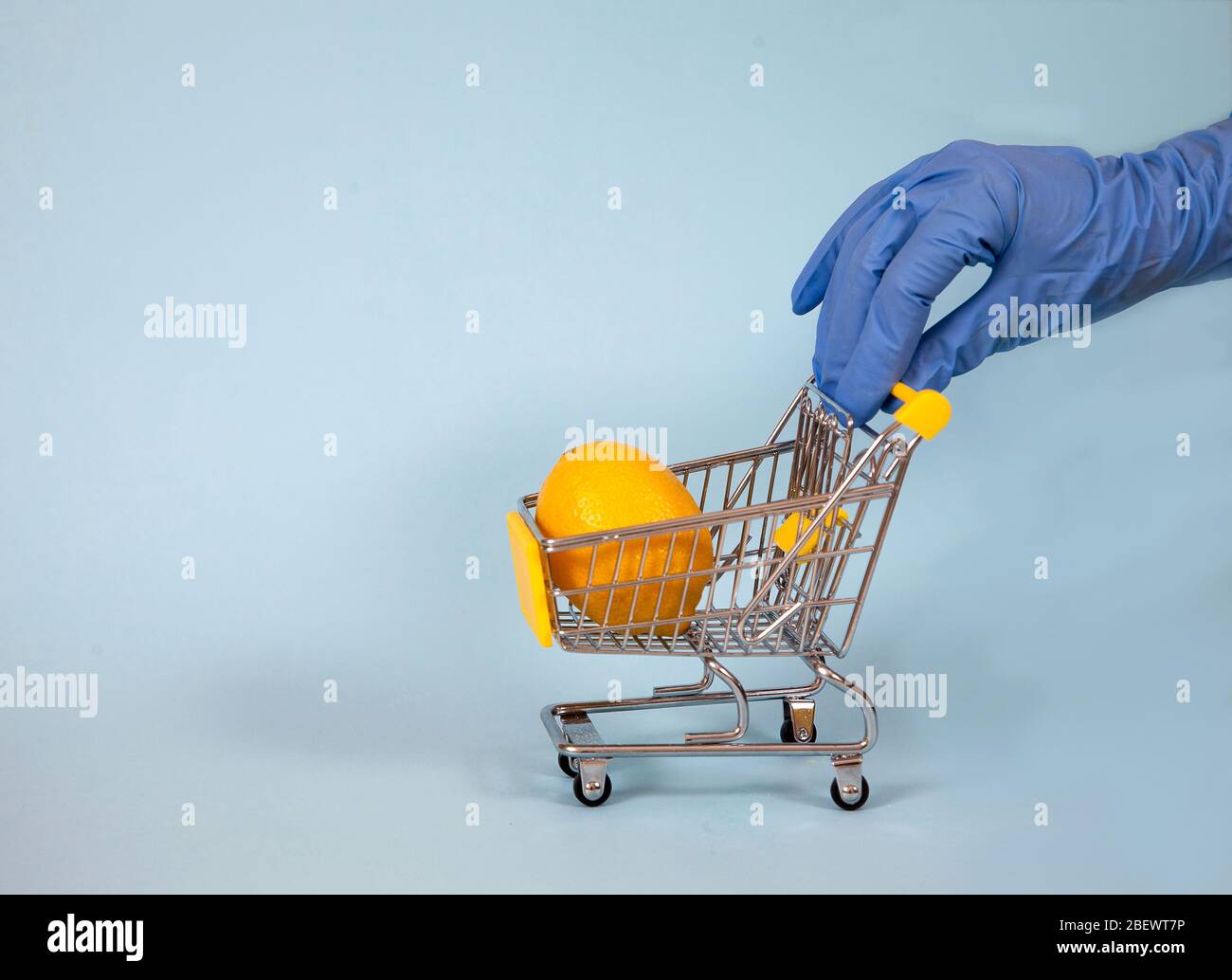 Mano femenina en un guante de goma azul enrolla un carrito de supermercado con limón sobre un fondo azul. Protección. Higiene. Covid 19 Foto de stock