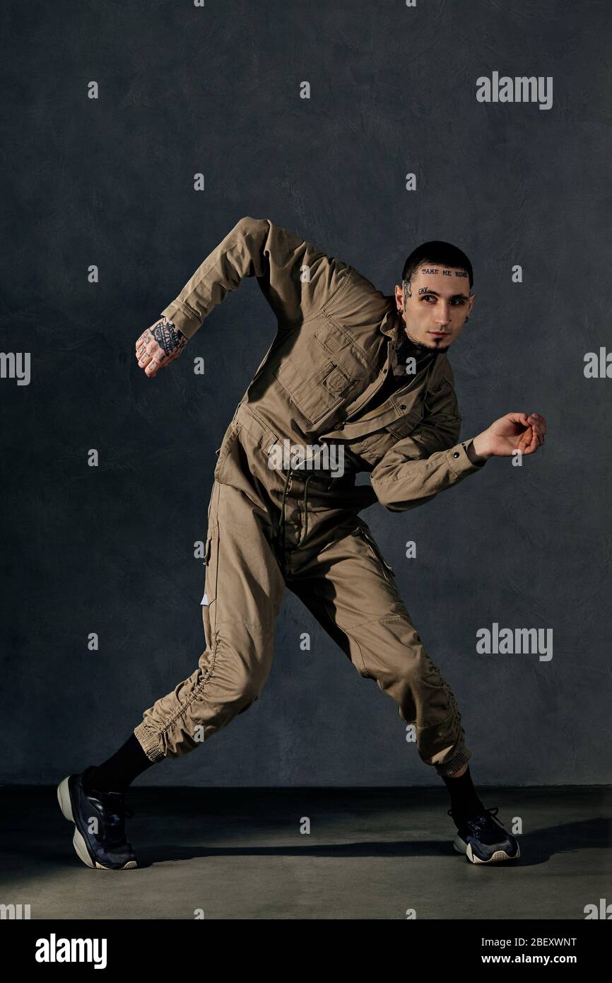 Joven con cuerpo y cara tatuados, pendientes, barba. Vestido con mono  caqui, sneakers negras. Baile sobre fondo gris. Salón de baile, hip-hop  Fotografía de stock - Alamy