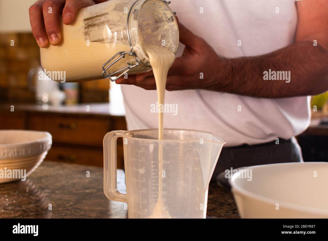 El pan de hornear del hombre mide el entrante de masa fermentada y vierte en una jarra mezcladora Foto de stock
