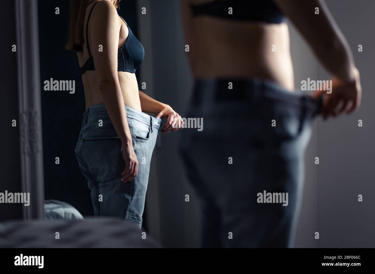 Mujer delgada y delgada vestida con pantalones grandes delante de un espejo después de la pérdida de peso y dieta. Niña con bajo peso con estrés malsano de ser gorda. Foto de stock