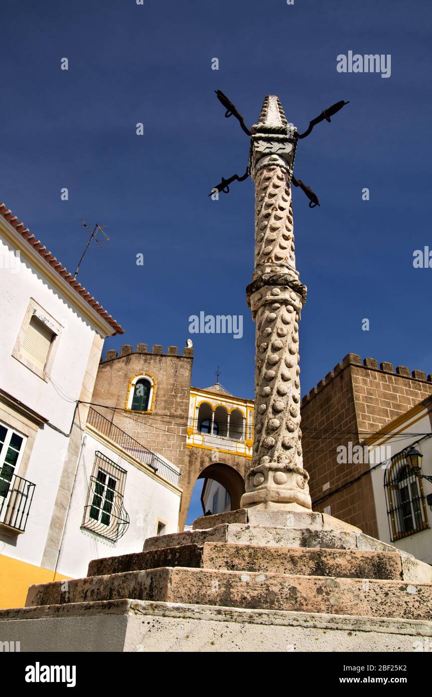 Antigua picota de piedra y hierro frente al arco de Santa Clara entre edificios de piedra y bajo un profundo cielo azul. Elvas, Portugal. Foto de stock