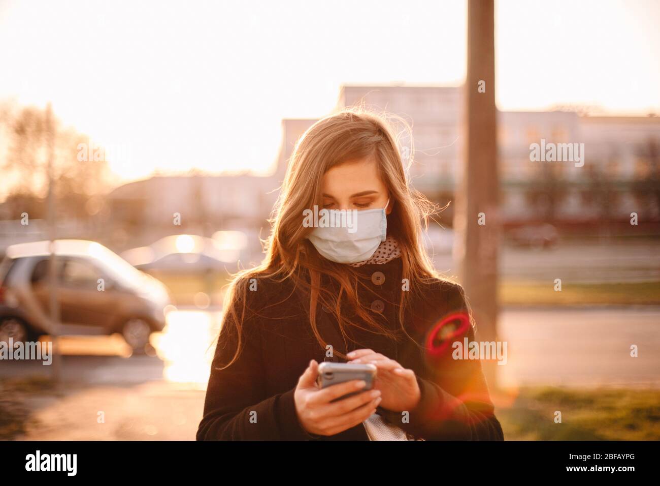 Mujer joven con máscara médica protectora facial mientras usa el teléfono inteligente caminando por la calle en la ciudad Foto de stock
