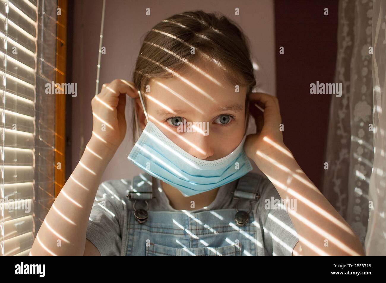 La niña está sentada en el alféizar de la ventana y tratando de poner una máscara desechable protectora en su cara durante el auto-aislamiento en casa debido al Coronavirus Foto de stock