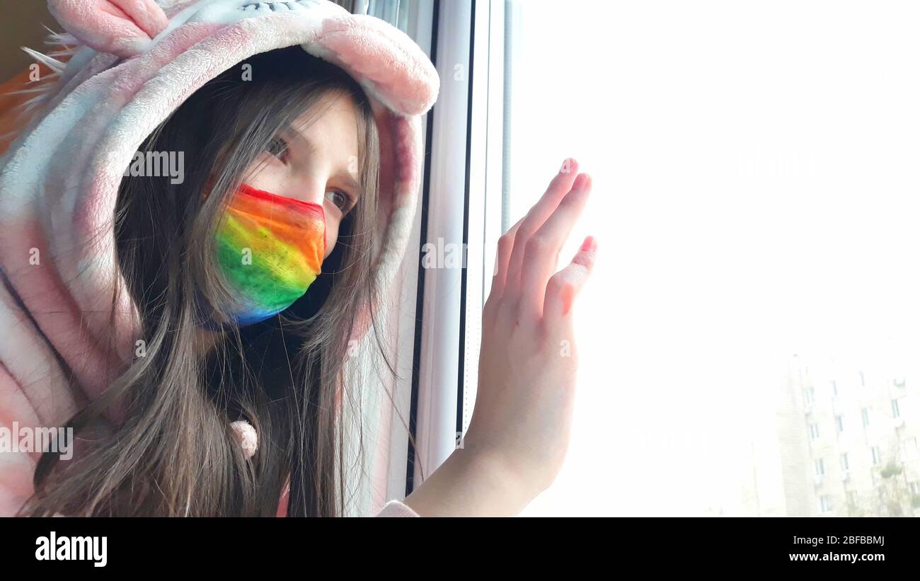 Una chica adolescente morena en una máscara médica pintada en colores brillantes arcoiris se encuentra en la ventana con su mano en glass.Concept de permanecer en casa, permaneciendo s Foto de stock