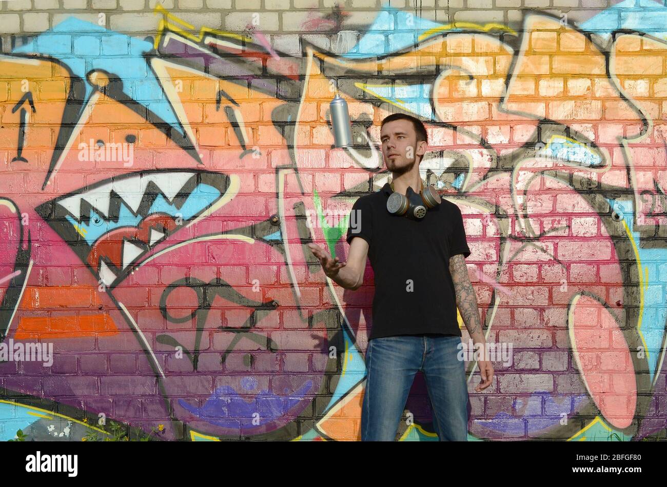 Joven artista de graffiti con máscara de gas en su cuello lanzar su aerosol  contra el colorido graffiti rosa en la pared de ladrillo. Arte callejero y  pintura contemporánea Fotografía de stock -