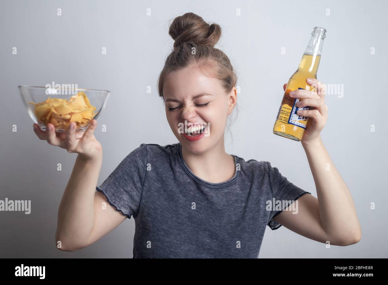 Sonriente joven mujer caucásica niña bebiendo una botella de cerveza y sosteniendo patatas fritas crujientes Foto de stock