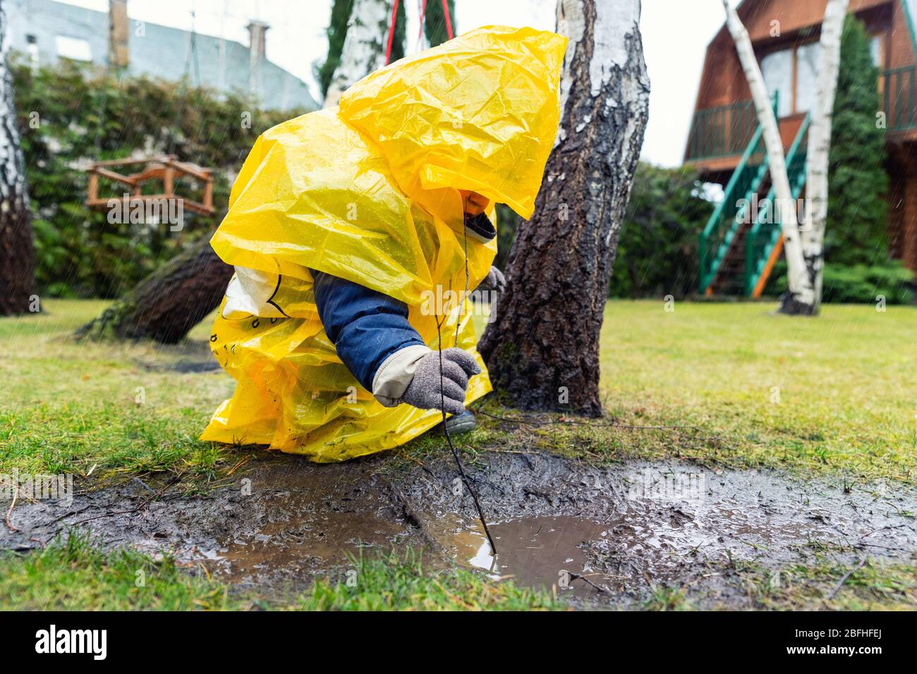 Lindo y adorable niño pequeño caucásico en un chubasquero amarillo  brillante y botas de agua jugando solo en un charco de barro durante el  frío día lluvioso de primavera en casa Fotografía