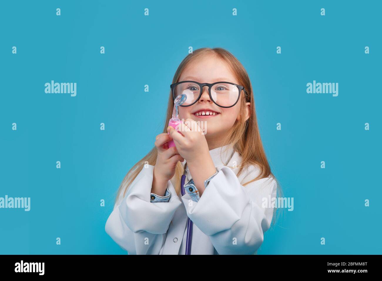 Niña sonriente con uniforme médico y gafas jugando con estetoscopio Foto de stock