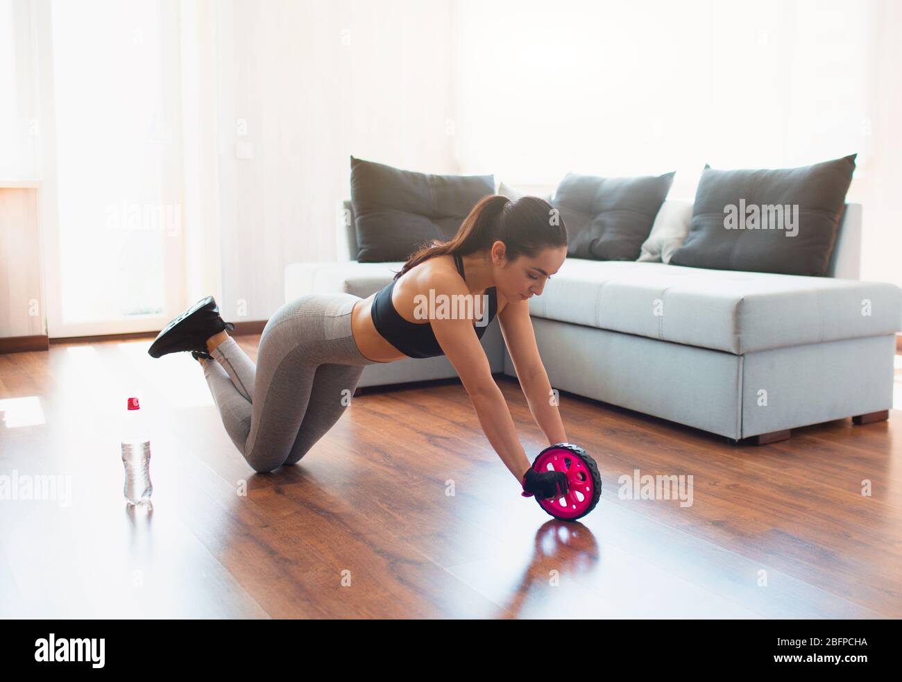 Mujer joven haciendo ejercicio deportivo en la habitación durante la  cuarentena. Póngase de pie sobre las rodillas y utilice el rodillo de  ejercicios abdominales para estirar el cuerpo. Formación en casa Fotografía
