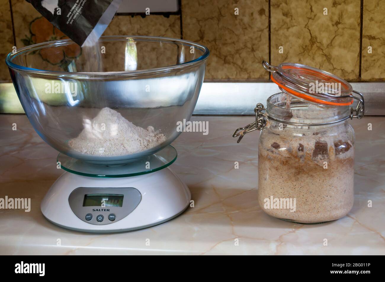 Pesando la harina lista para hacer pan de masa fermentada con el arrancador de masa fermentada activado en un tarro de cristal para horno. Foto de stock