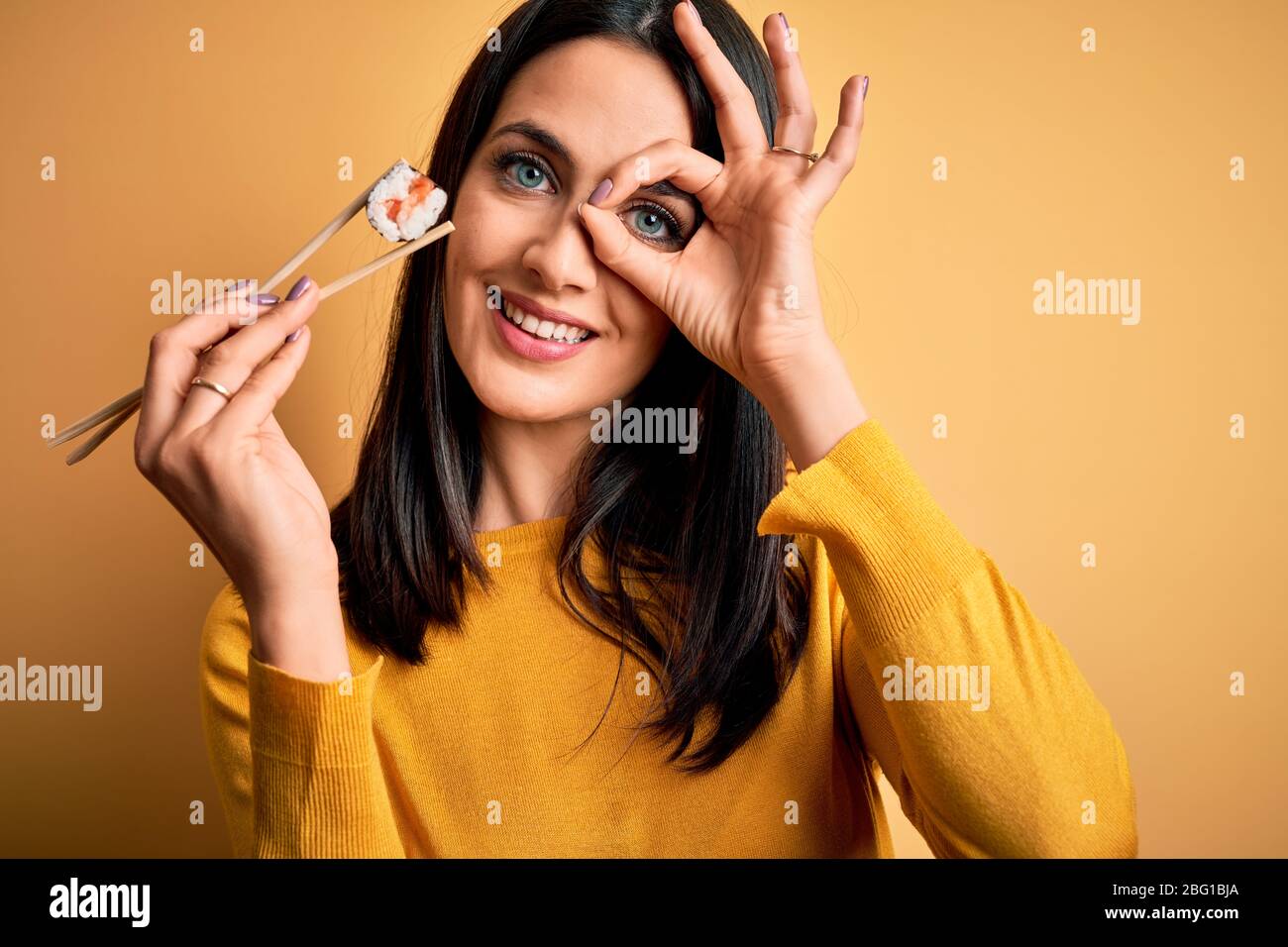 Joven mujer morena con ojos azules comiendo salmón maki sushi usando  palillos con cara feliz sonriendo haciendo ok signo con la mano en el ojo  mirando a través de f Fotografía de