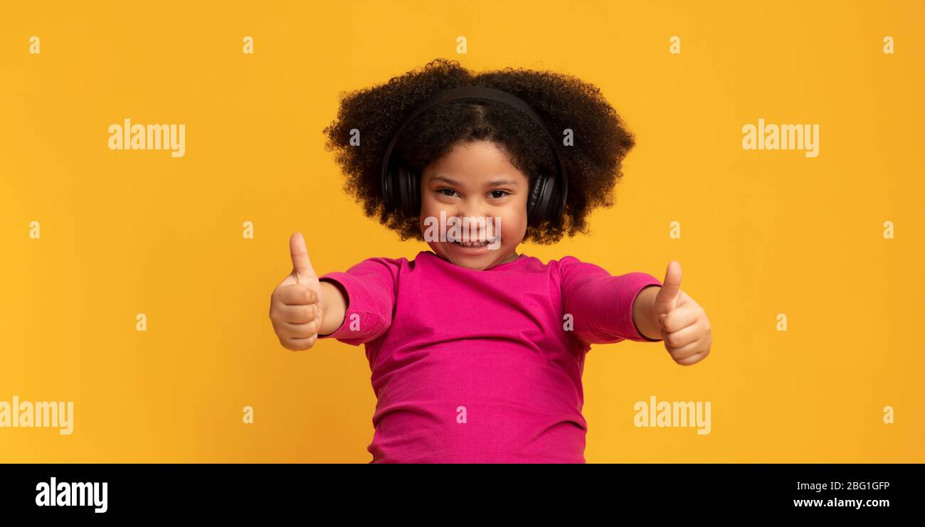 Una chica pequeña y positiva que escucha música en los auriculares inalámbricos y muestra el pulgar hacia arriba Foto de stock