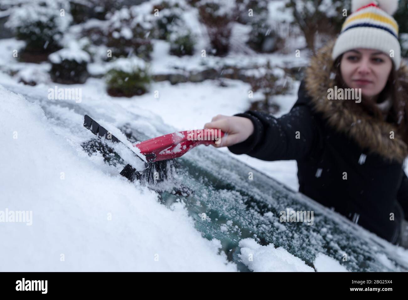 Joven mujer morena atractiva limpiando la nieve de un coche con un raspador de hielo en el invierno. Foto de stock