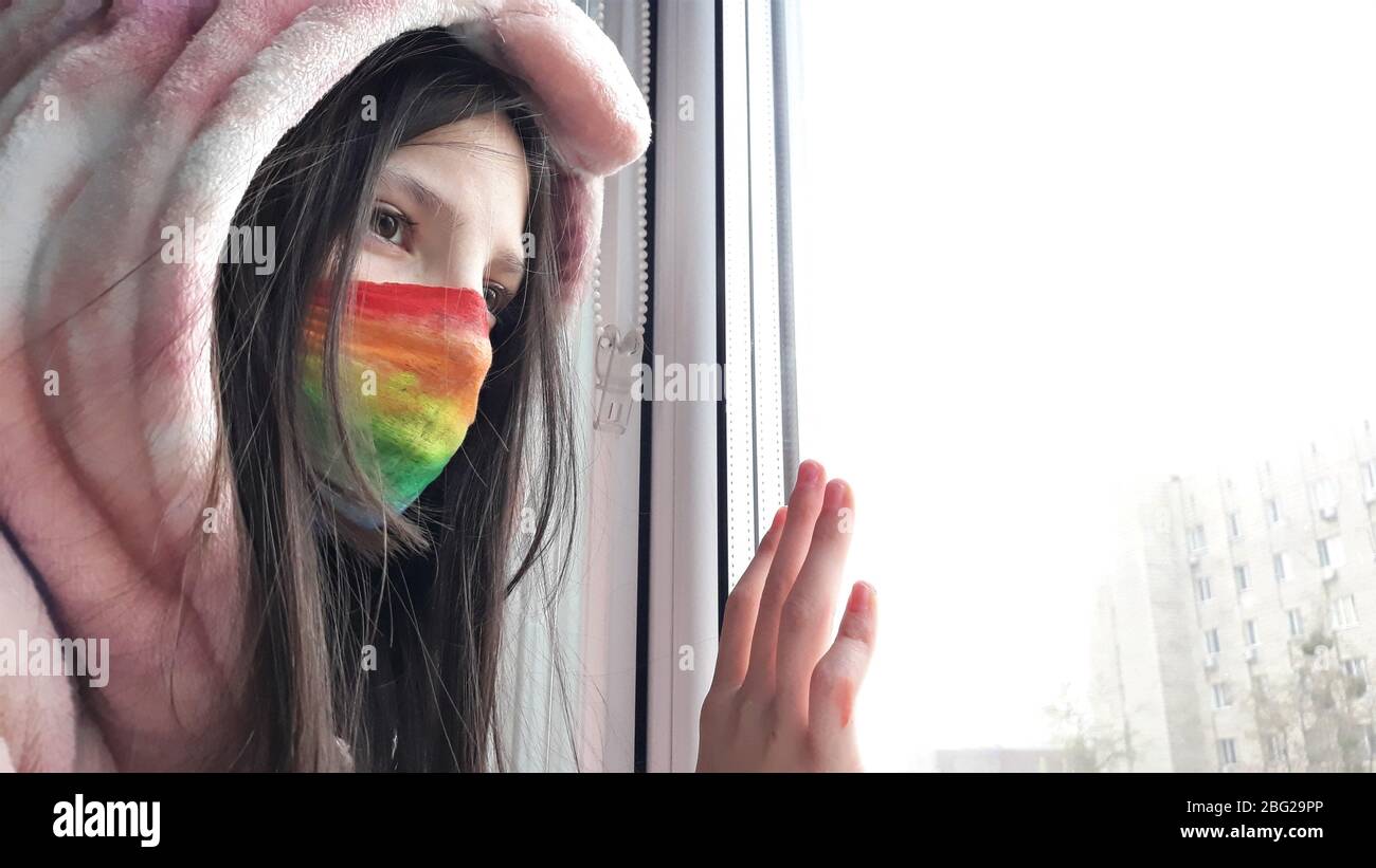 Una chica adolescente morena en una máscara médica pintada en colores brillantes arcoiris se encuentra en la ventana con su mano en glass.Concept de permanecer en casa, quedándose Foto de stock