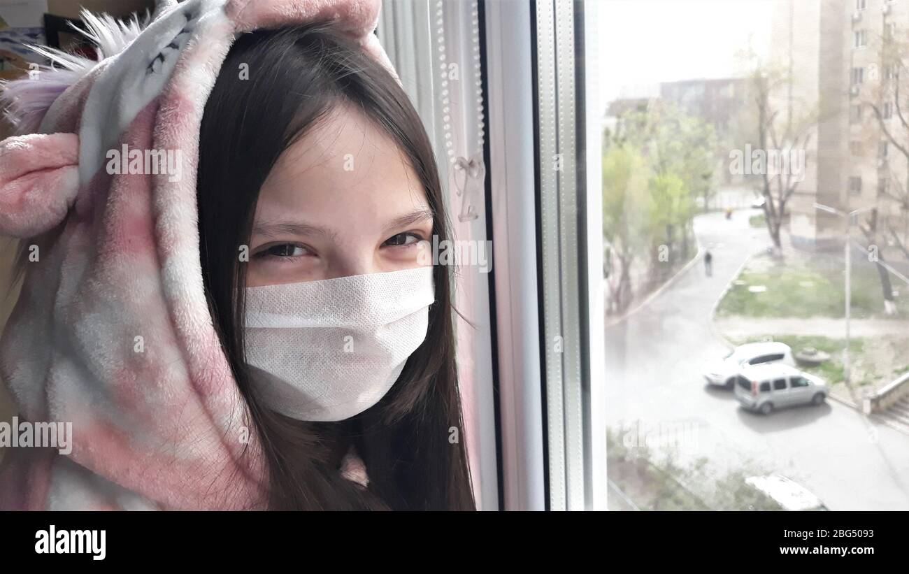 Una chica adolescente morena en una máscara médica se encuentra en la ventana con su mano en glass.Concept de permanecer en casa, permanecer seguro.aprendizaje a distancia Foto de stock