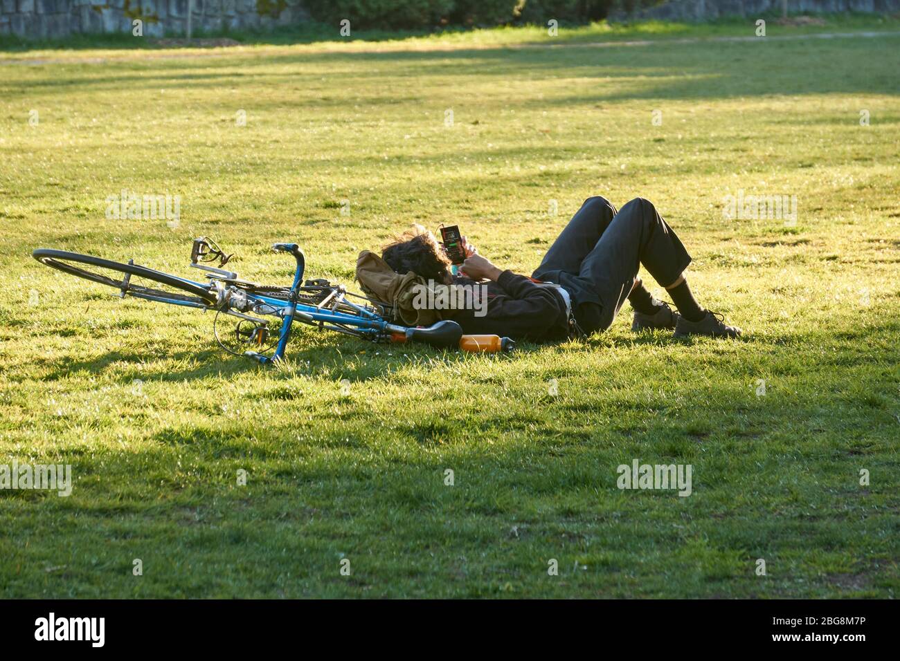 Vancouver, Canadá, 18 de abril de 2020. Un joven se encuentra solo en la hierba utilizando su dispositivo electrónico para permanecer conectado durante la pandemia del COVD-19. Foto de stock