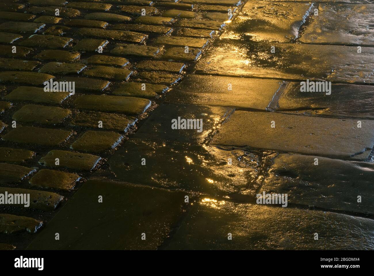 Vista baja del pavimento mojado por la noche mostrando losas y bloques redondeados más pequeños con iluminación de la calle reflejada en la humedad Foto de stock