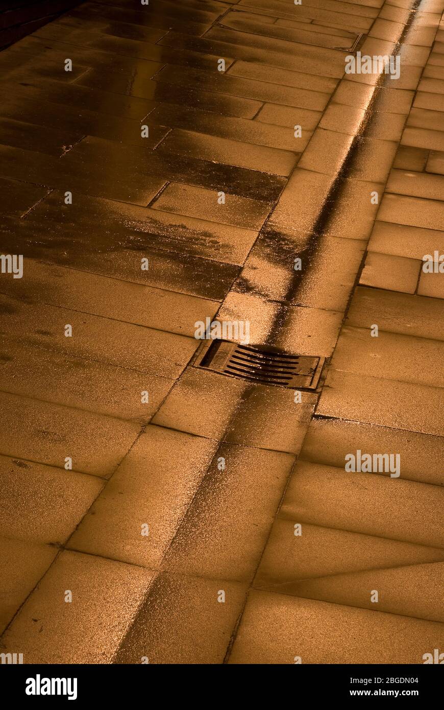 Banderas de pavimentación húmeda con canal central, desagüe y parrilla, todo con un brillo dorado de la iluminación reflejada de la calle Foto de stock
