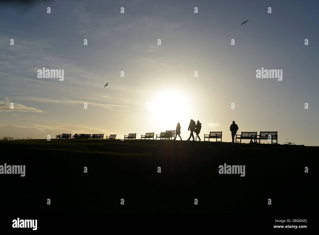 El sol que se hunde crea una silueta sorprendente de cuatro figuras que caminan por una fila de asientos de banco en una colina con efecto de sol de halo y cielo azul pálido Foto de stock