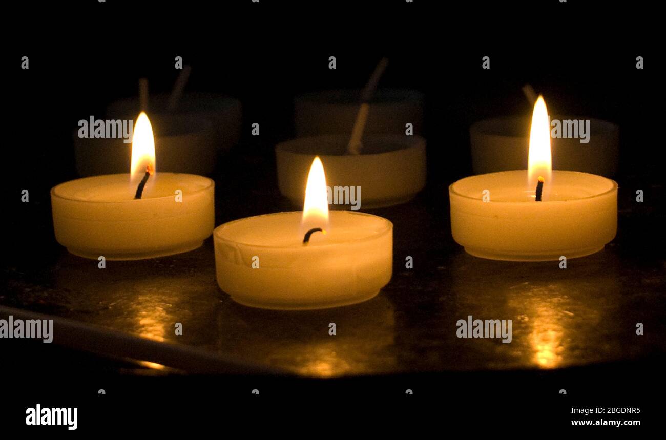 Vista de cerca de tres velas votivas encendidas en un soporte con otras sin iluminación detrás y desvaneciéndose en el fondo Foto de stock