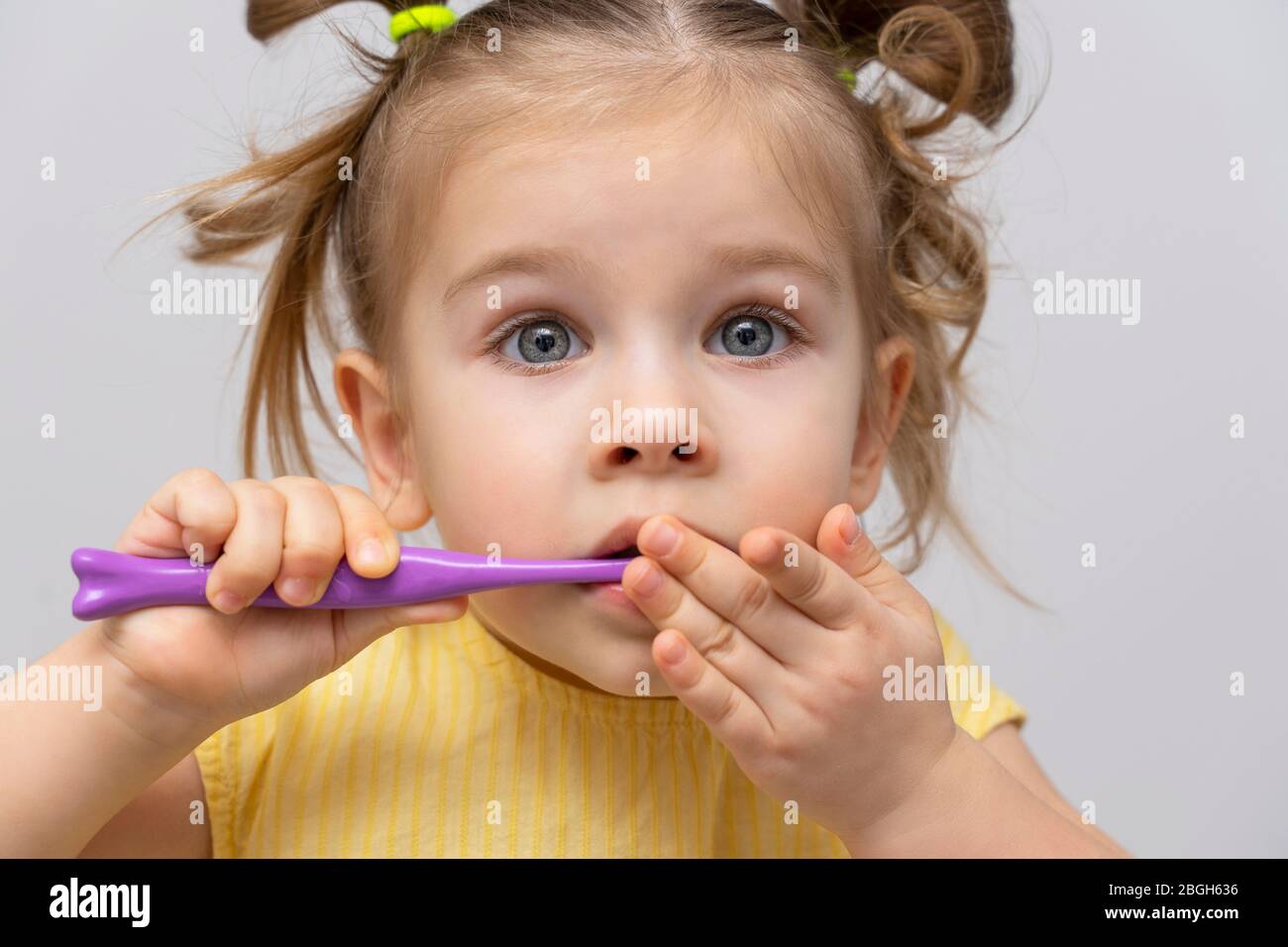 una niña pequeña en una camisa amarilla se está cepillando los dientes limpios y cubre la boca con su mano. emocional con los ojos asustados. problemas con los dientes y las encías Foto de stock