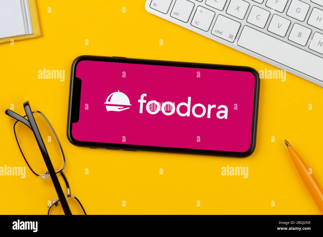 Un smartphone que muestra el logotipo de Foodora descansa sobre un fondo amarillo junto con un teclado, gafas, lápiz y libro (sólo para uso editorial). Foto de stock