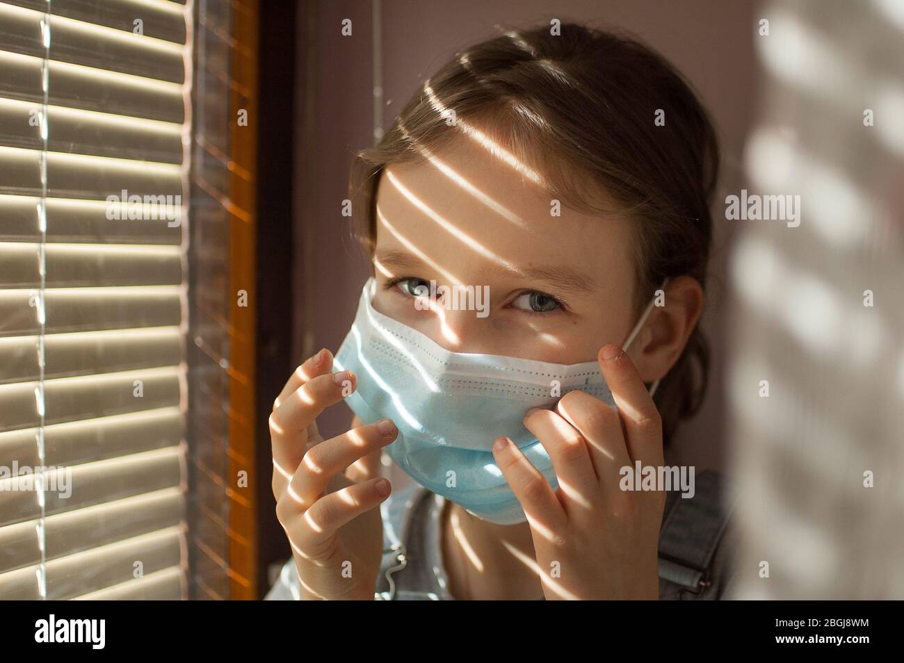 La niña está sentada en el alféizar de la ventana y tratando de poner una máscara desechable protectora en su cara durante el auto-aislamiento en casa debido al Coronavirus Foto de stock