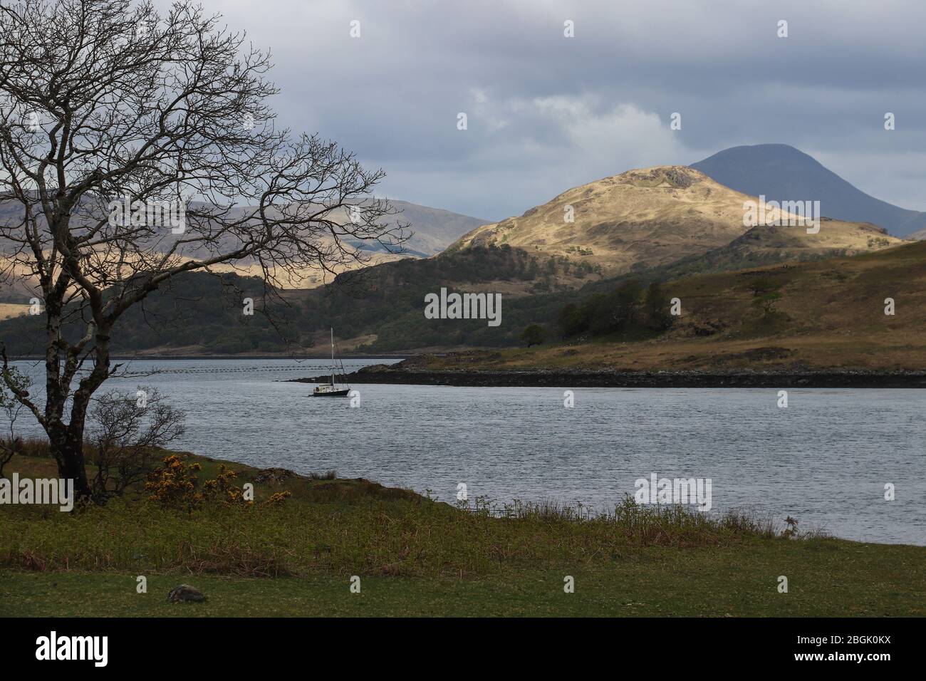 Un solitario velero sube al lago Spelve en la isla de Mull en Escocia. Un parche de luz solar ilumina la colina cercana, Ben azul más en la distancia Foto de stock