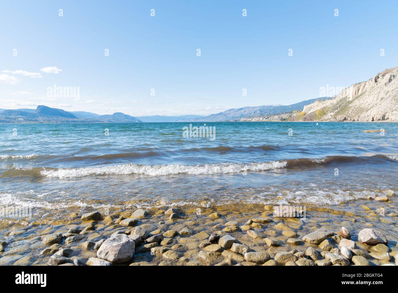 Olas rompiendo en la costa rocosa del lago Okanagan con vista al cielo azul y a la distante cordillera Foto de stock