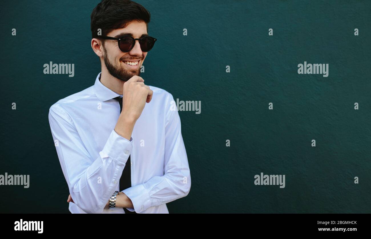 hombre de negocios en gafas de sol mirando hacia otro lado y sonriendo con su mano en la barbilla. Hombre caucásico joven de pie contra una pared oscura con espacio de copia. Foto de stock