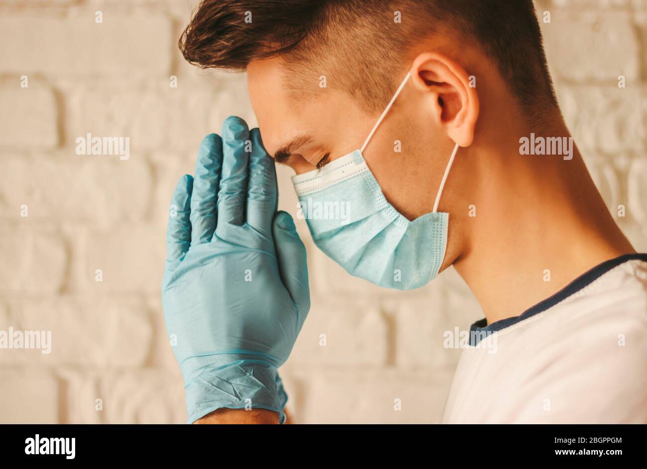 Médico religioso con guantes protectores y máscara médica rezando por ayudar a Dios en el hospital. Oración de joven enfermo en guantes médicos en manos y prote Foto de stock