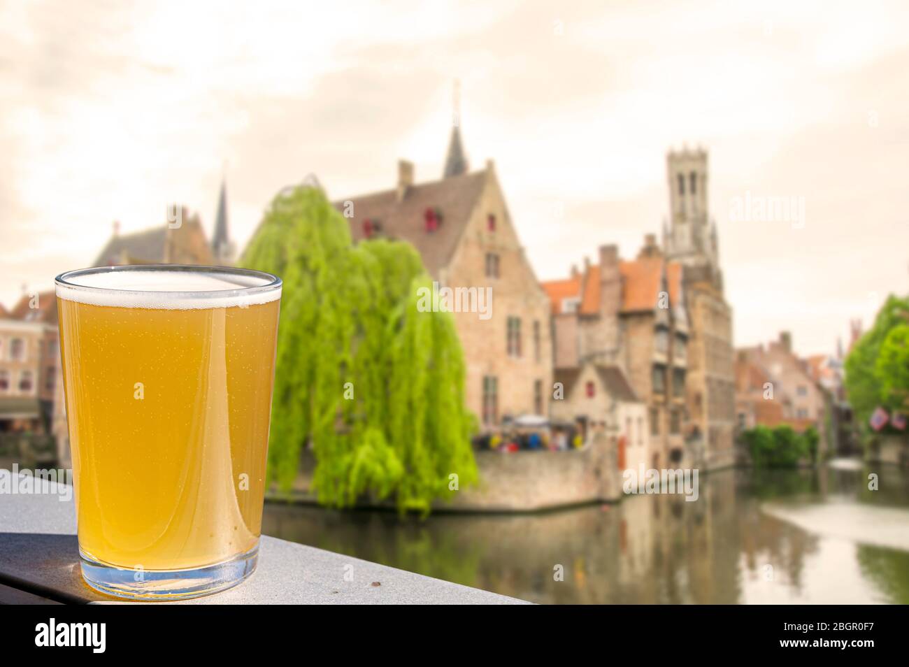 Copa de cerveza con vistas a casas históricas, canal y torre del campanario en el centro de la ciudad de Brujas, Bélgica. Foto de stock