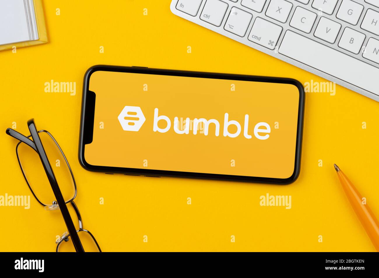 Un smartphone que muestra el logotipo de Bumble descansa sobre un fondo amarillo junto con un teclado, gafas, lápiz y libro (sólo para uso editorial). Foto de stock