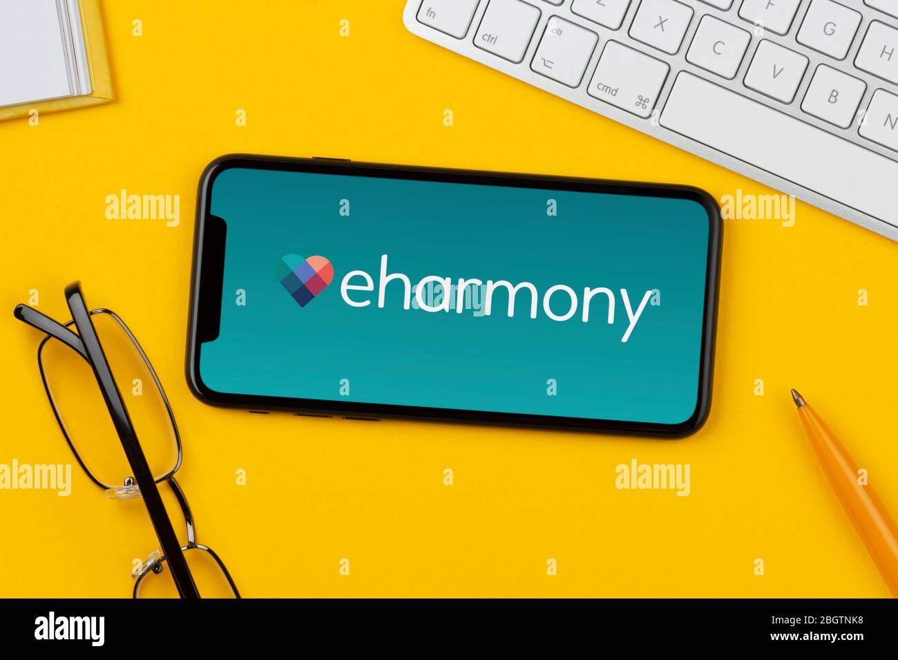 Un smartphone que muestra el logotipo de eHarmony descansa sobre un fondo amarillo junto con un teclado, gafas, lápiz y libro (sólo para uso editorial). Foto de stock