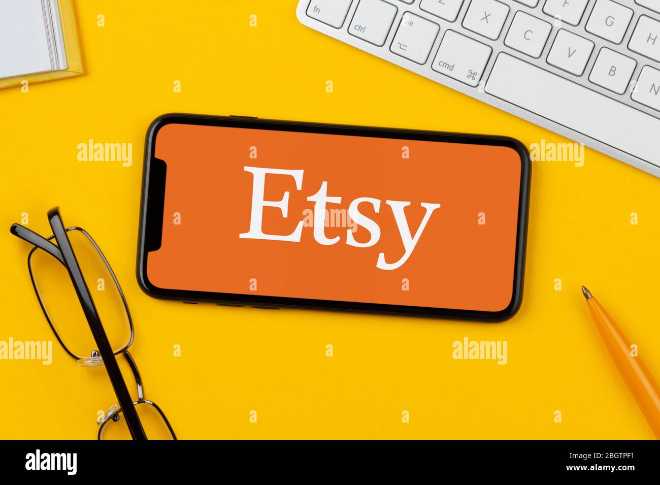 Un smartphone que muestra el logotipo de Etsy descansa sobre un fondo amarillo junto con un teclado, gafas, lápiz y libro (sólo para uso editorial). Foto de stock