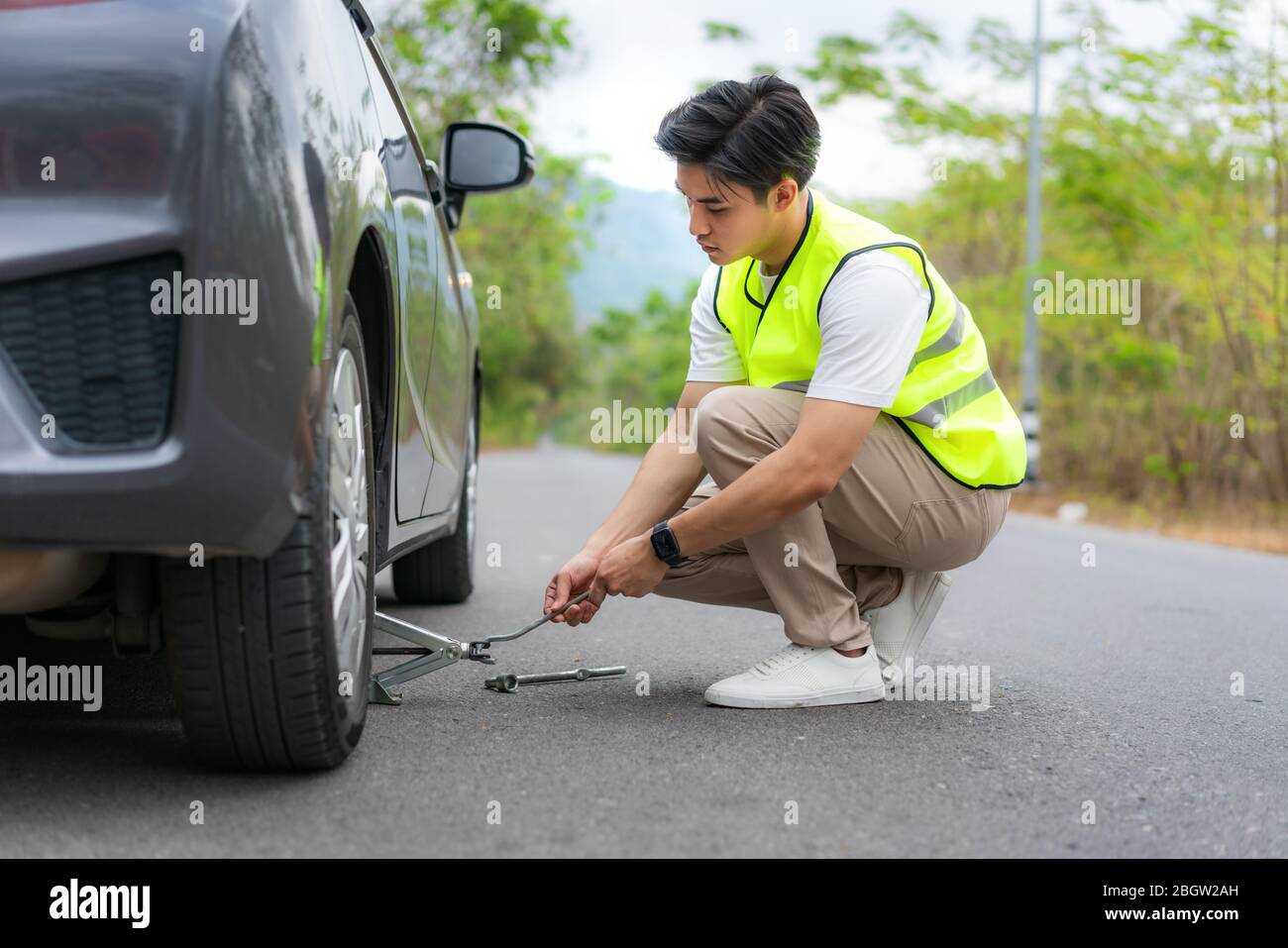 Hombre asiático joven con chaleco verde de seguridad cambiando el neumático perforado en su coche aflojando las tuercas con una llave de rueda antes de levantar el vehículo con el gato Foto de stock